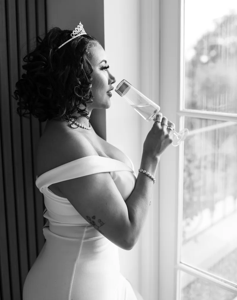 Black and white portrait of the bride sipping champagne by the window