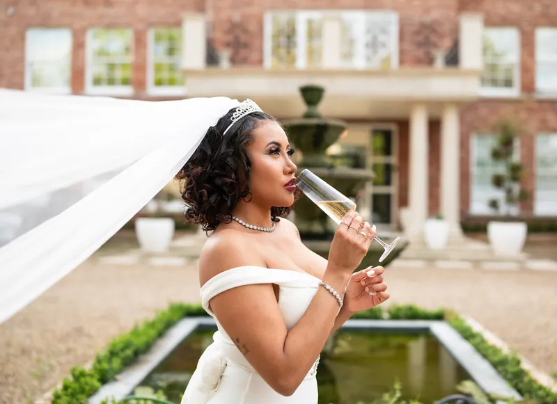 Bride sipping champagne by the pond as her veil blows in the wind
