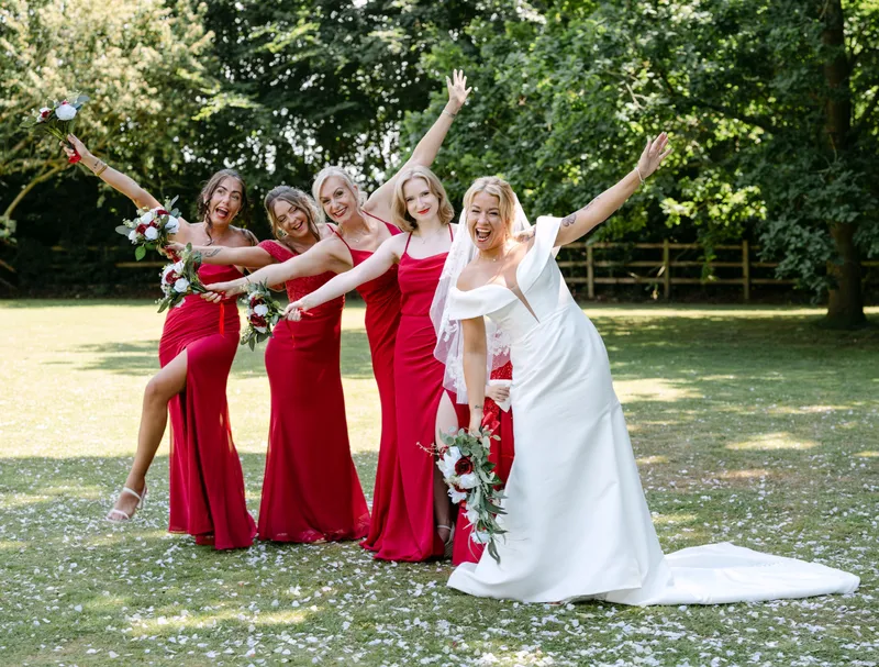 Bride and bridesmaids in red dresses cheering on the lawn at a Kent wedding