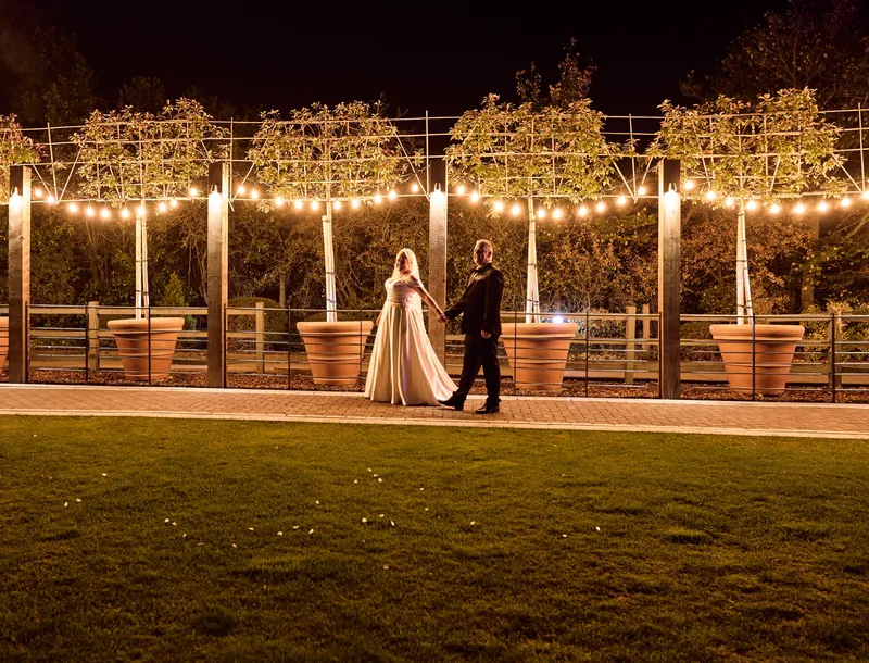 Bride and groom walking under festoon lights and pleached trees at night