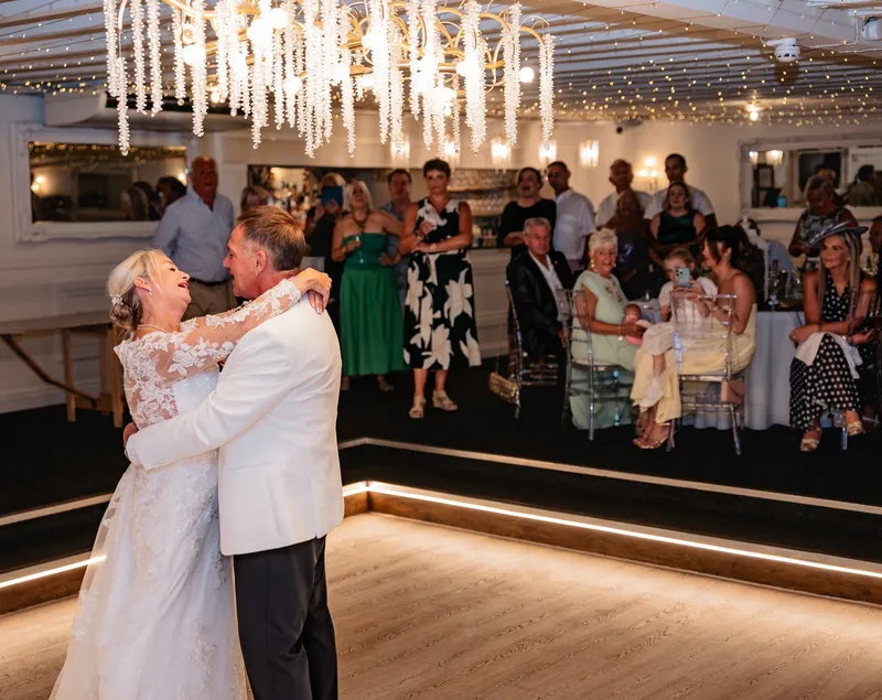 Bride laughing up at the groom during the first dance under a crystal chandelier as guests watch