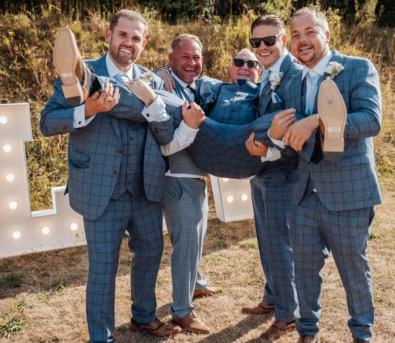 Groomsmen in matching check suits carrying the groom in the sunshine