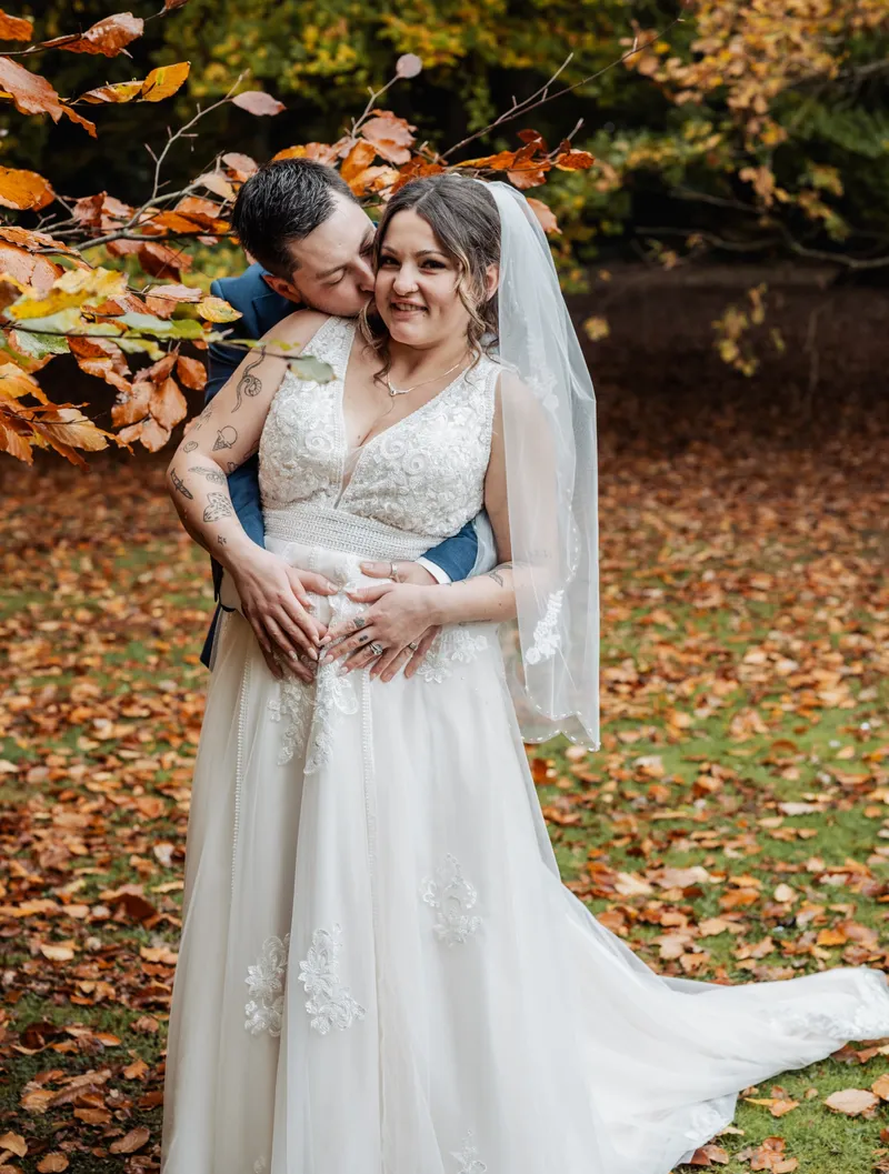 Groom hugging the bride from behind under autumn leaves at Oakwood House