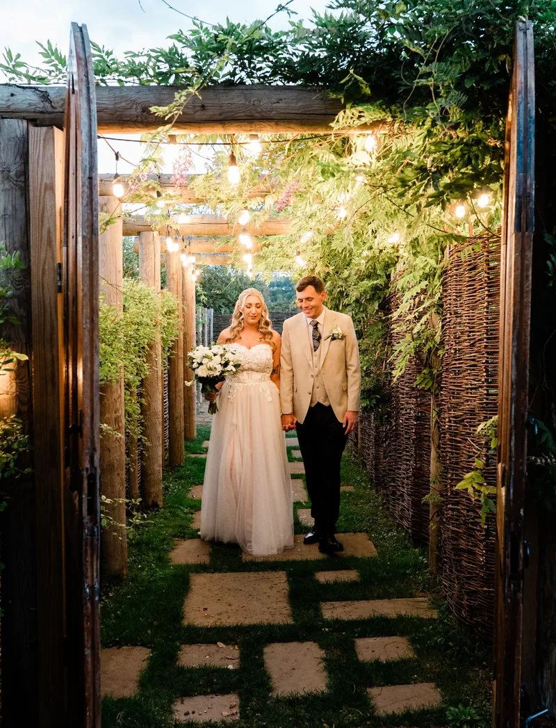 Couple walking through wooden gates under a lit wisteria pergola at The Orchard