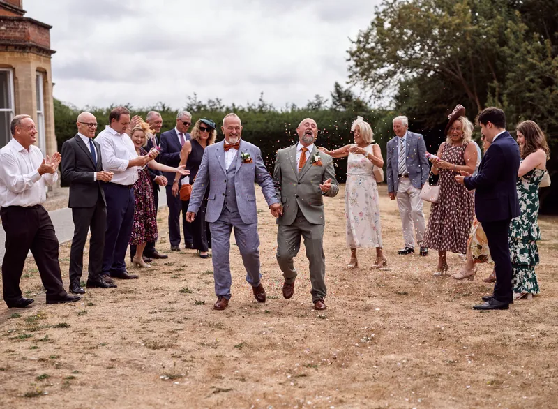 Two grooms laughing as guests throw confetti outside a Kent wedding venue