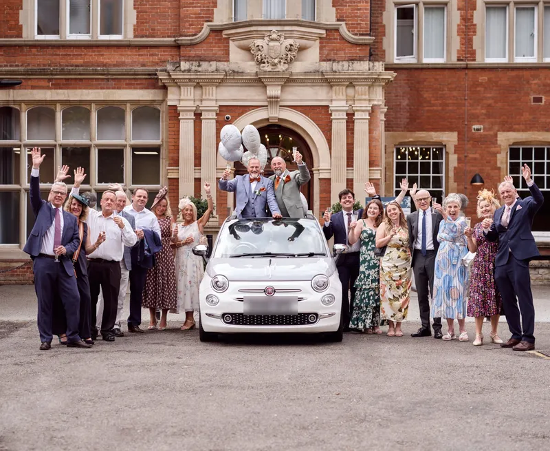 Two grooms raising champagne through a car sunroof as guests cheer outside a Kent wedding venue